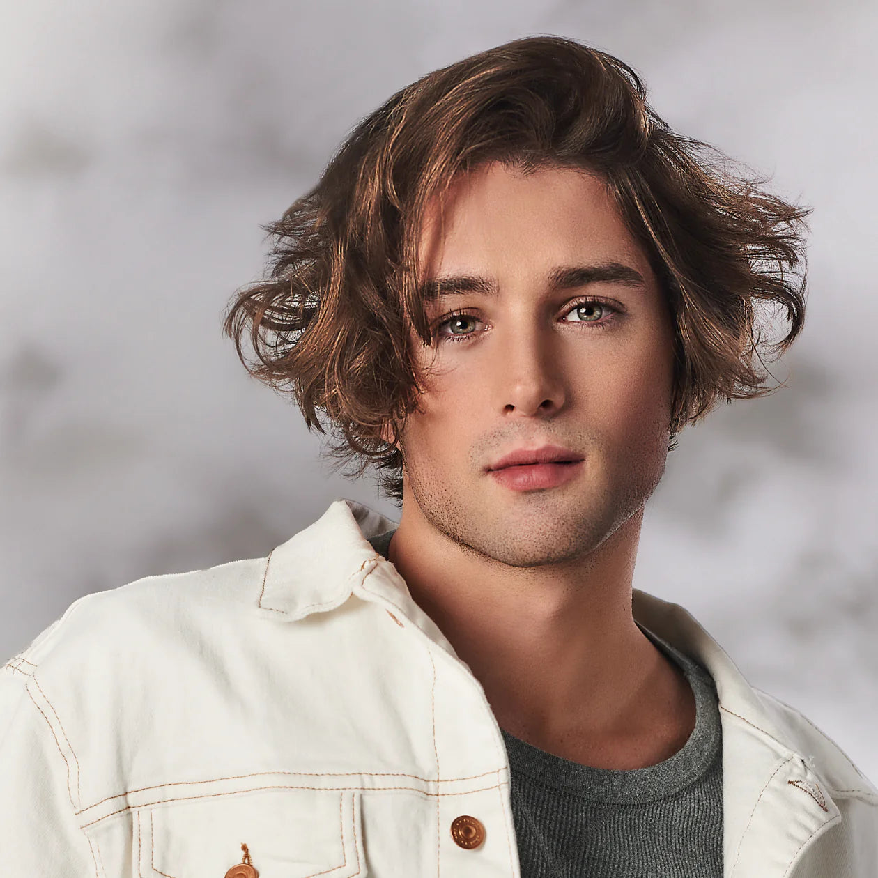 Brunette model with short, wavy, loose curls in a white denim jacket in front of a blurred gray background.