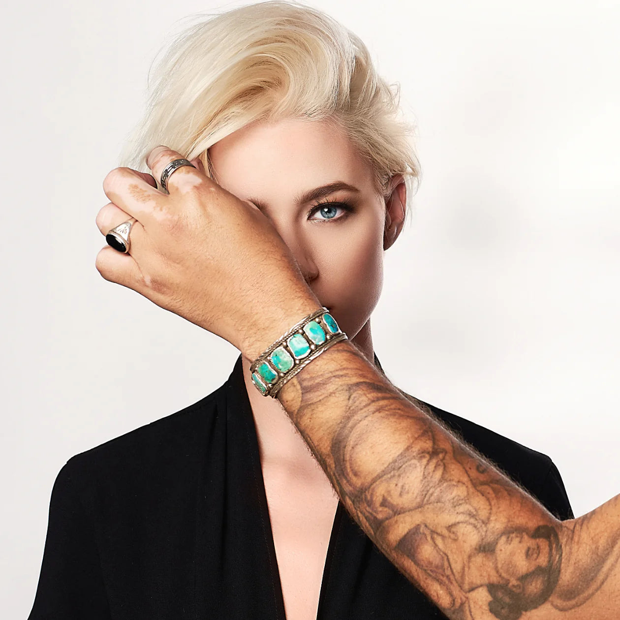 Blonde model with a sleek hairstyle gazes at the camera as a tattooed hand, adorned with a turquoise bracelet, styles her hair against a white backdrop, highlighting the vibrant jewelry and artistic tattoos